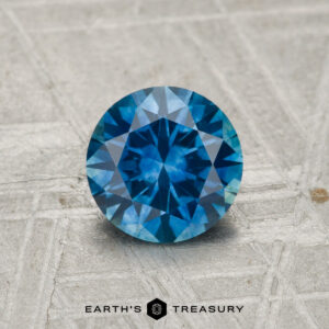 A round, faceted 1.07-carat heated Montana Sapphire with a blue hue rests on a textured, light surface, with "Earth's Treasury" visible below.
