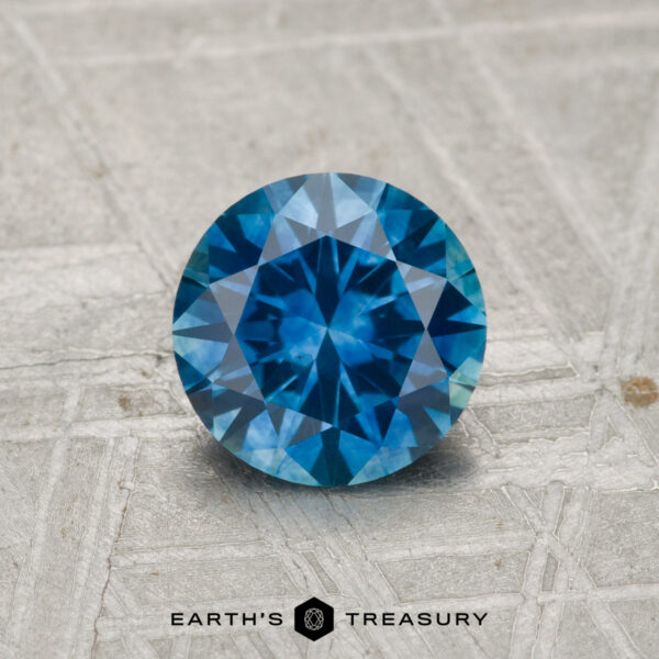 1.07-Carat Montana Sapphire (Heated) A round, faceted 1.07-carat heated Montana Sapphire with a blue hue rests on a textured, light surface, with "Earth's Treasury" visible below.
