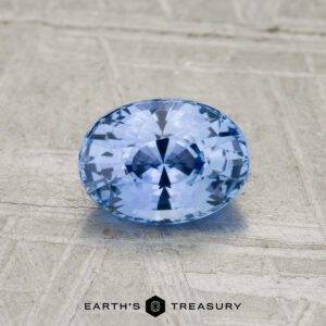 1.11-carat Ceylon sapphire, oval-cut and blue, displayed on a textured gray surface with "EARTH'S TREASURY" text at the bottom.