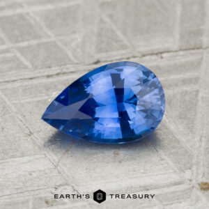 1.12-Carat Ceylon Sapphire in a pear shape, blue hue, shown on a textured gray surface with the "Earth's Treasury" logo at the bottom.
