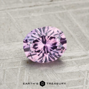 A 1.09-carat, oval-cut Montana sapphire with a light purple hue is displayed on a textured gray surface, with "EARTH'S TREASURY" written below.