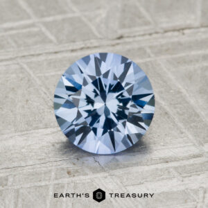A 1.37-carat Ceylon sapphire, round brilliant-cut and blue, is displayed on a textured gray surface with the text "Earth's Treasury" below.
