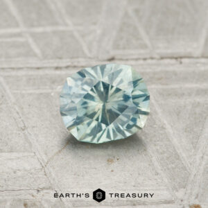 A 0.71-carat Montana sapphire, faceted in an oval shape with a light blue-green hue, is shown on a textured gray surface with "EARTH'S TREASURY" at the bottom.