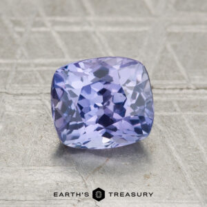 A faceted, cushion-cut 1.63-carat Ceylon Sapphire with a bluish-purple hue is displayed on a textured metallic surface. The text "Earth's Treasury" appears at the bottom.