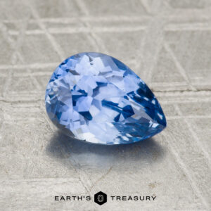 A 1.55-carat Ceylon sapphire, cut in a pear shape with facets, is displayed on a textured surface with "EARTH'S TREASURY" written below.