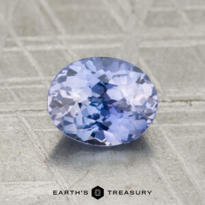 A 1.41-carat Ceylon sapphire, oval-cut and light blue, rests on a textured metallic surface with "EARTH'S TREASURY" displayed below.