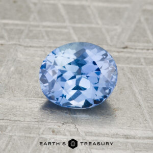A 1.35-carat Ceylon sapphire, faceted in an oval shape, rests on a textured gray surface viewed from above. "EARTH'S TREASURY" appears at the bottom of the image.