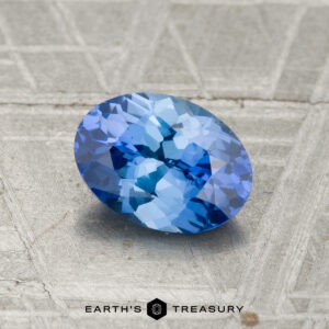 A 1.17-carat Ceylon sapphire with an oval cut and faceted surface rests on a textured gray background labeled "EARTH'S TREASURY" at the bottom.