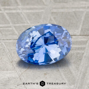 A 1.72-carat Ceylon Sapphire, oval-cut, rests on a textured gray surface with "EARTH'S TREASURY" and a hexagonal logo at the bottom.