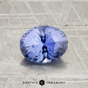A 1.48-carat Ceylon Sapphire in a faceted oval cut rests on a textured gray surface, with "Earth's Treasury" displayed at the bottom.