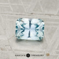 A 1.04-carat, rectangular faceted Montana Sapphire in light blue rests on a textured grey surface. "EARTH'S TREASURY" is visible at the bottom of the image.