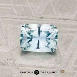 A 1.04-carat, rectangular faceted Montana Sapphire in light blue rests on a textured grey surface. "EARTH'S TREASURY" is visible at the bottom of the image.