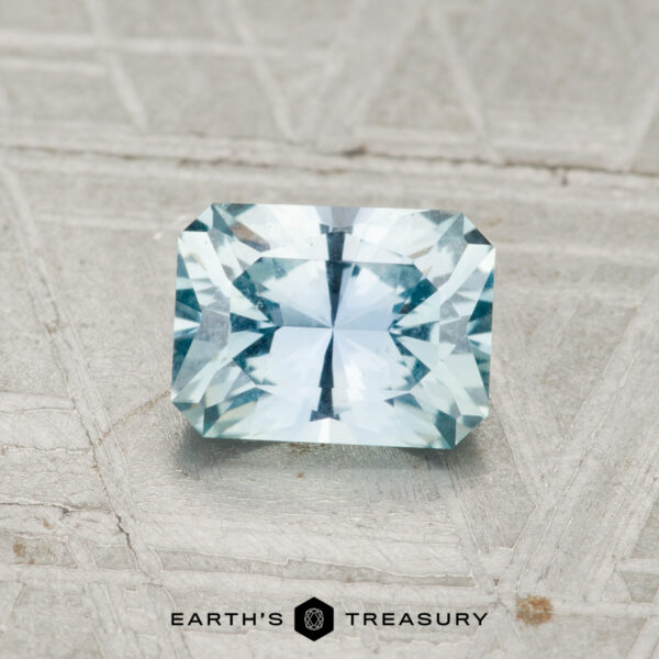 1.04-Carat Montana Sapphire A 1.04-carat, rectangular faceted Montana Sapphire in light blue rests on a textured grey surface. "EARTH'S TREASURY" is visible at the bottom of the image.