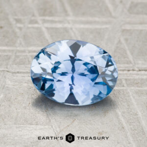 A 1.57-carat Ceylon sapphire with an oval cut is displayed on a textured gray surface, with the text "EARTH'S TREASURY" shown below.