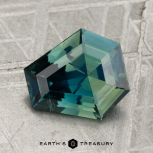 A 2.44-carat heated Ceylon sapphire with deep blue-green geometric facets rests on a textured gray surface; the Earth's Treasury logo is displayed below.