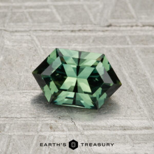 A 1.26-carat faceted hexagonal green sapphire rests on a textured gray surface with the "EARTH'S TREASURY" text and logo at the bottom of the image.