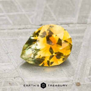 A faceted, pear-shaped 1.44-carat Montana sapphire in yellow and green hues is displayed on a textured gray surface with "EARTH'S TREASURY" text below.