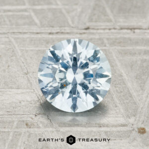 A 1.40-carat Montana sapphire in a round brilliant cut is displayed on a textured metallic surface with "EARTH'S TREASURY" at the bottom.