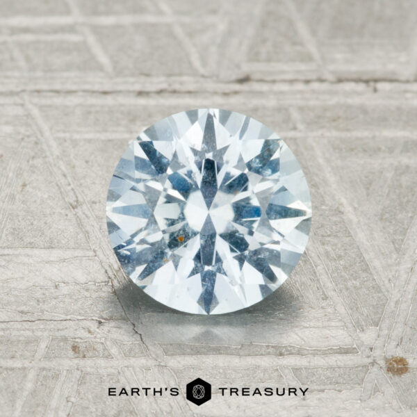 1.40-Carat Montana Sapphire A 1.40-carat Montana sapphire in a round brilliant cut is displayed on a textured metallic surface with "EARTH'S TREASURY" at the bottom.