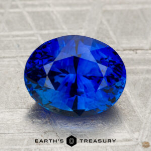 A 3.67-carat heated Ceylon sapphire, oval-cut and deep blue, displayed on a textured gray surface with the text "Earth's Treasury" below.