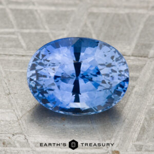 A 3.06-carat heated Ceylon sapphire is displayed on a textured metallic surface labeled "Earth's Treasury.