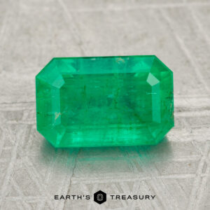 A 2.25-carat rectangular-cut green emerald gemstone rests on a textured gray surface with "EARTH'S TREASURY" displayed at the bottom.