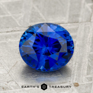 A 2.18-carat heated Ceylon sapphire with round faceting is displayed on a textured gray surface, with "EARTH'S TREASURY" printed below.