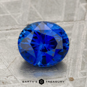 A 2.18-carat heated Ceylon sapphire with round faceting is displayed on a textured gray surface, with "EARTH'S TREASURY" printed below.