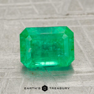 A 1.94-carat emerald-cut green gemstone is showcased on a textured gray surface with "EARTH'S TREASURY" text beneath it.