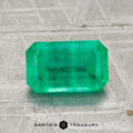 A 1.59-carat rectangular faceted green emerald gemstone is shown on a textured gray surface with "EARTH'S TREASURY" written below.