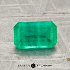 A 1.59-carat rectangular faceted green emerald gemstone is shown on a textured gray surface with "EARTH'S TREASURY" written below.