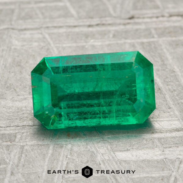 1.56-Carat Emerald A 1.56-carat rectangular-cut green emerald gemstone sits on a textured gray surface, with the "Earth's Treasury" logo displayed at the bottom.