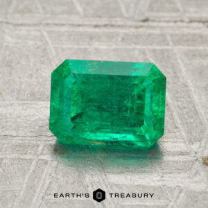 A 1.53-carat rectangular-cut green emerald is displayed on a light textured surface with the text "EARTH'S TREASURY" at the bottom.