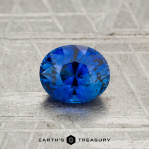 A 1.04-carat heated Ceylon sapphire, cut in a faceted oval, is showcased on a textured gray surface with "EARTH'S TREASURY" at the bottom.