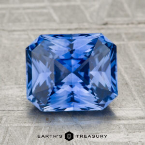 A 2.72-carat heated Ceylon sapphire with a faceted rectangular cut is displayed on a gray textured surface, featuring the "EARTH'S TREASURY" text below.