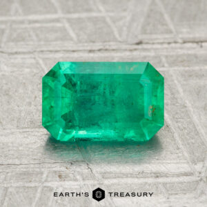 A 1.40-carat rectangular, faceted green emerald rests on a textured gray surface. "EARTH'S TREASURY" appears at the bottom of the image.