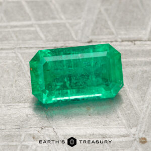 A 1.39-carat emerald-cut green gemstone rests on a textured metallic surface, with the logo and text "EARTH'S TREASURY" below.
