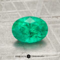 A 1.38-carat oval-cut green emerald rests on a textured gray surface with the "Earth's Treasury" logo and text at the bottom.