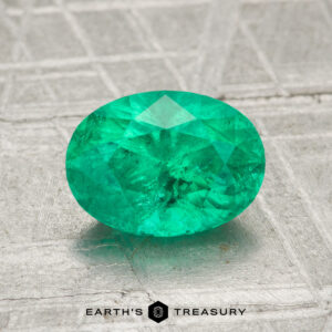 A 1.38-carat oval-cut green emerald rests on a textured gray surface with the "Earth's Treasury" logo and text at the bottom.