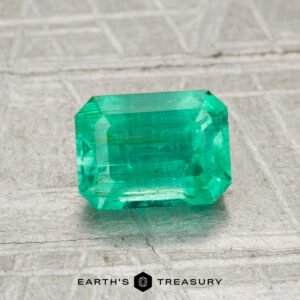 A 1.13-carat rectangular cut green emerald is shown on a textured gray surface with "EARTH'S TREASURY" at the bottom.