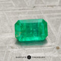 A 1.00-carat emerald with a rectangular cut is placed on a textured gray surface, featuring the text "EARTH'S TREASURY" at the bottom.