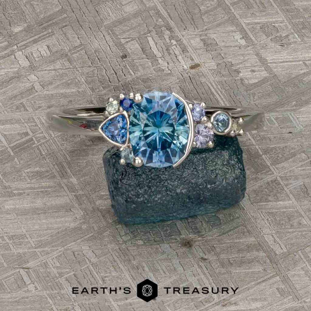 Mielke, Christine [custom gallery]: Silver ring with blue gemstone center and small accent stones, displayed on rough blue crystal; 'Earth’s Treasury' logo below.