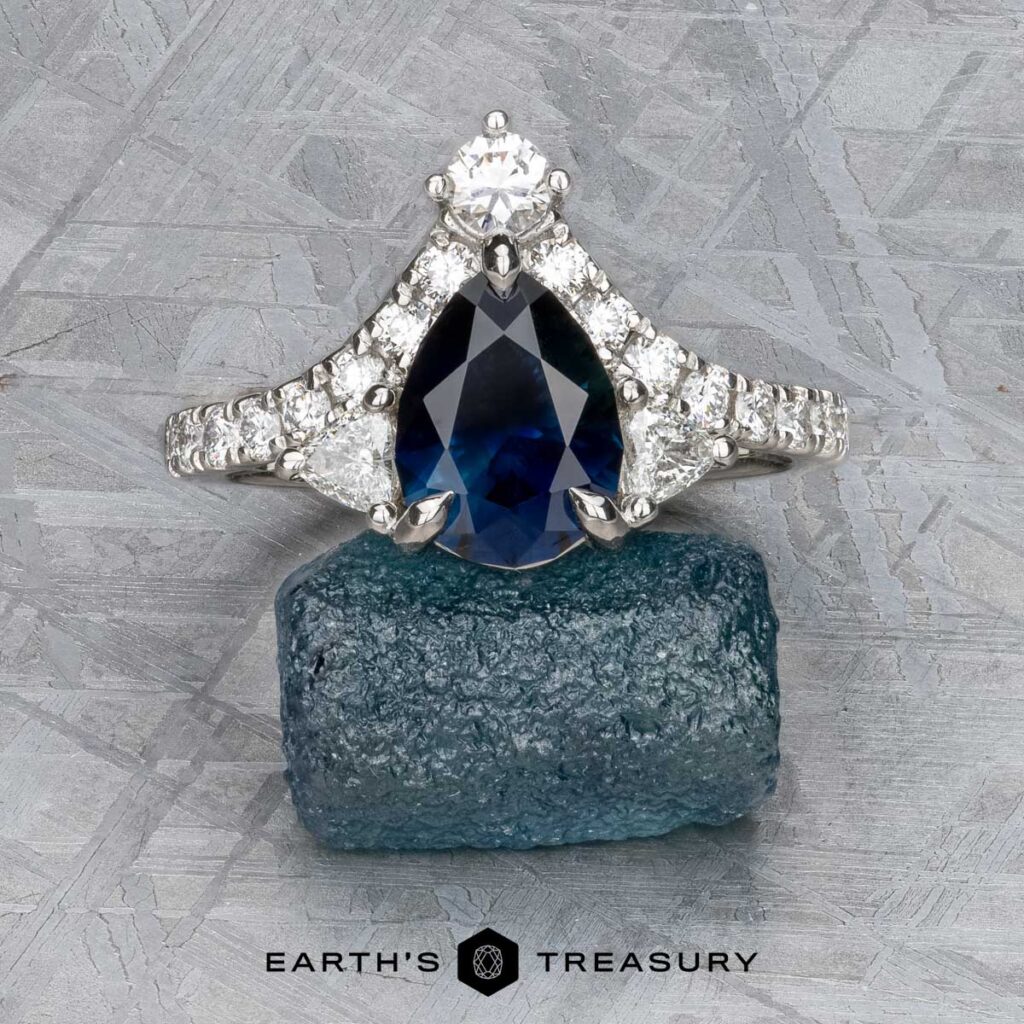 A pear-shaped blue sapphire and diamond ring set in white metal, displayed on a rough blue gemstone, with "Connolly, Sean and Martirano, Julia 1 [custom gallery]" below.