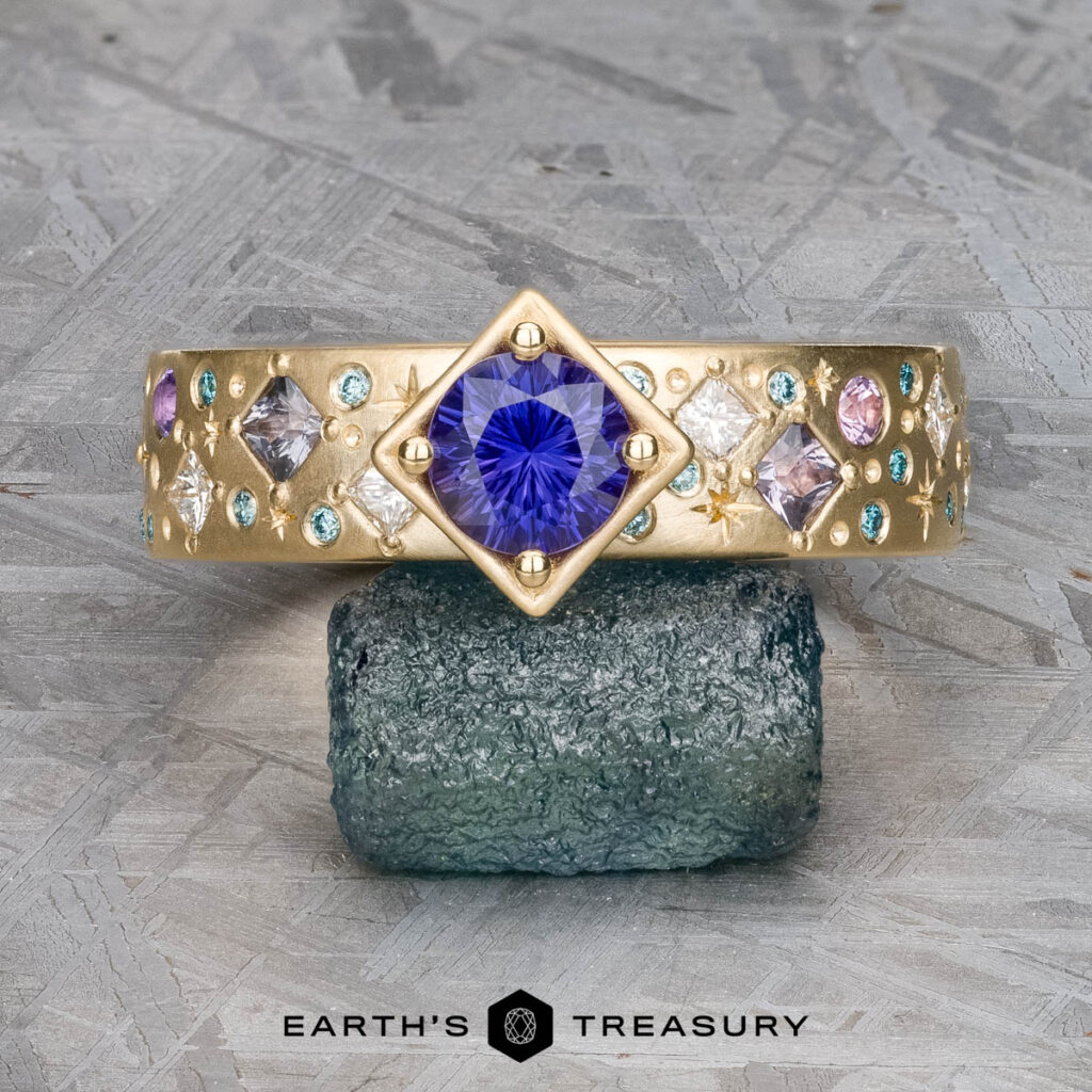 The "Mielke, Christine 7 [custom gallery]" gold ring features a central blue gemstone in a square bezel, encircled by smaller colored stones and displayed on a rough green stone.