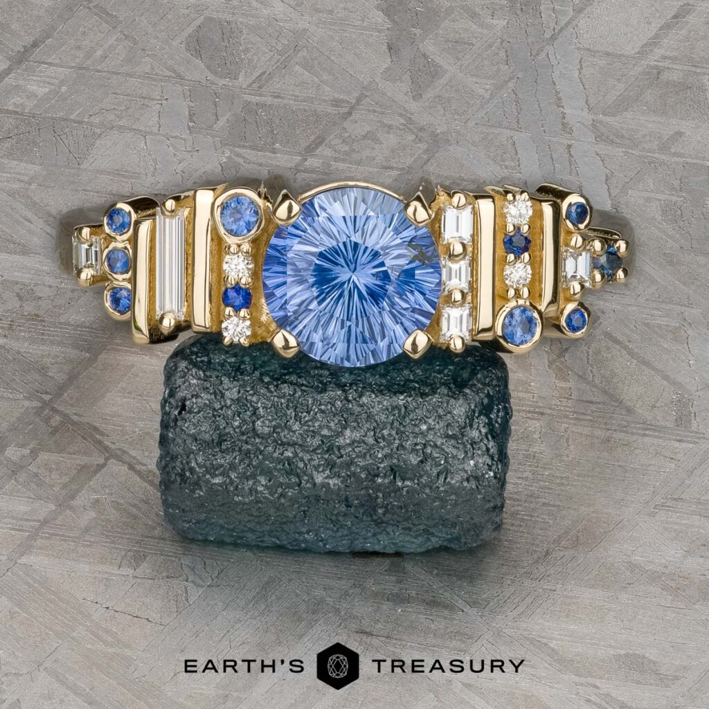 The Mielke, Christine [custom gallery] features a gold ring with a large round blue centerpiece, accented by smaller blue and clear stones, showcased on a rugged dark stone background.