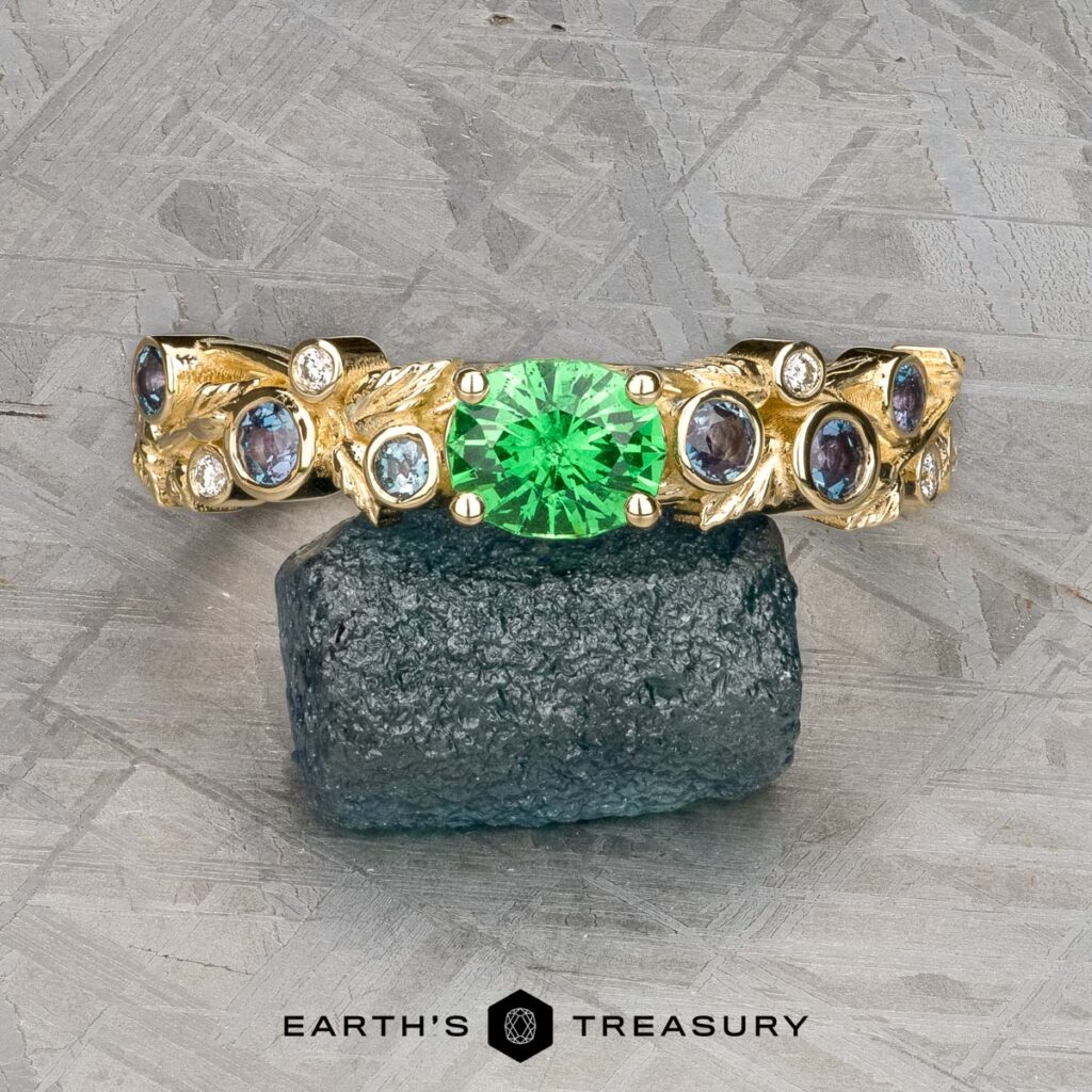 Mielke, Christine [custom gallery] gold ring featuring a green gemstone center with blue and clear accents, displayed on a dark mineral, 'Earth's Treasury' logo beneath.