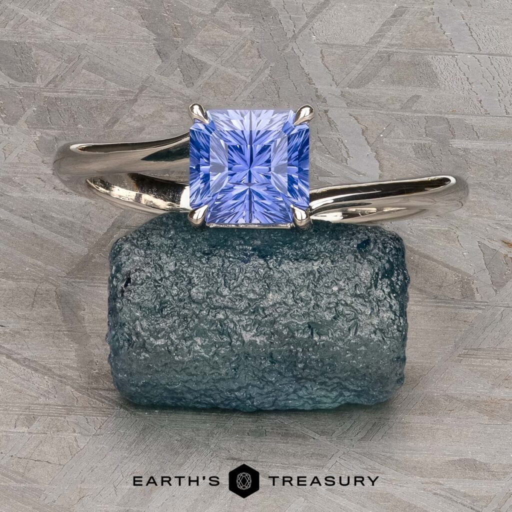 The Fiamor, Jonathan silver ring with a square-cut blue gemstone is showcased on a rough blue stone, set atop a textured gray surface.