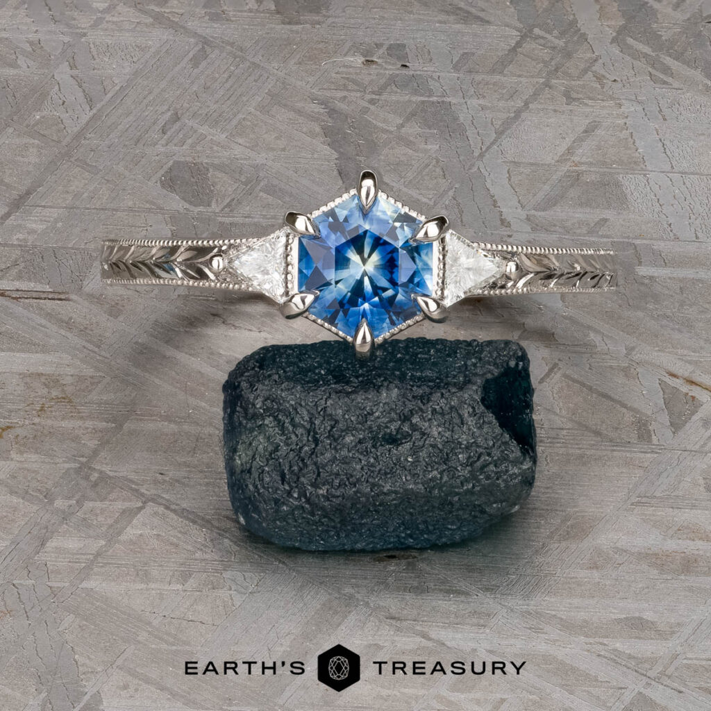The McMahon, Mary and Jamie [custom gallery] silver ring features a hexagonal blue gemstone and two triangular clear stones, showcased balanced on a dark rough stone with the "Earth's Treasury" logo below.
