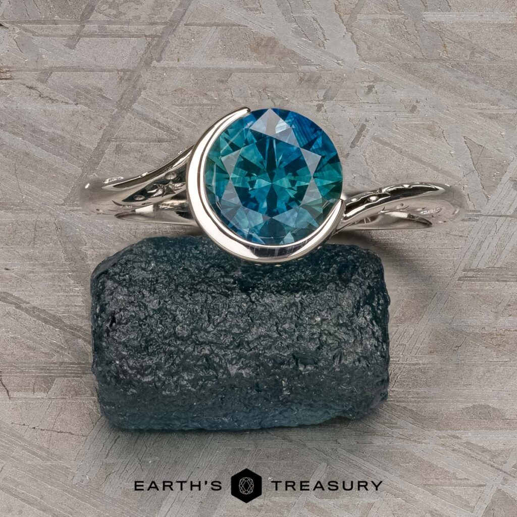 The "Hirani, Hanisha [custom gallery]" silver ring with a round blue gemstone rests on a rough dark stone against a textured gray background. The "Earth's Treasury" logo is shown below.