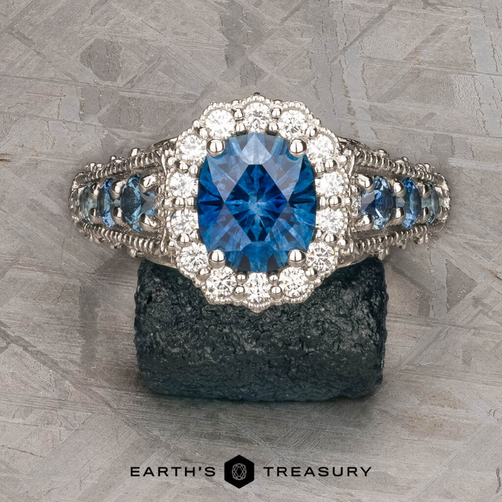 The Hailer, Sarah (Beth) and Zurawski, Jason custom gallery features a silver ring with a large oval blue gemstone at the center, surrounded by smaller clear stones and displayed on a dark mineral rock.